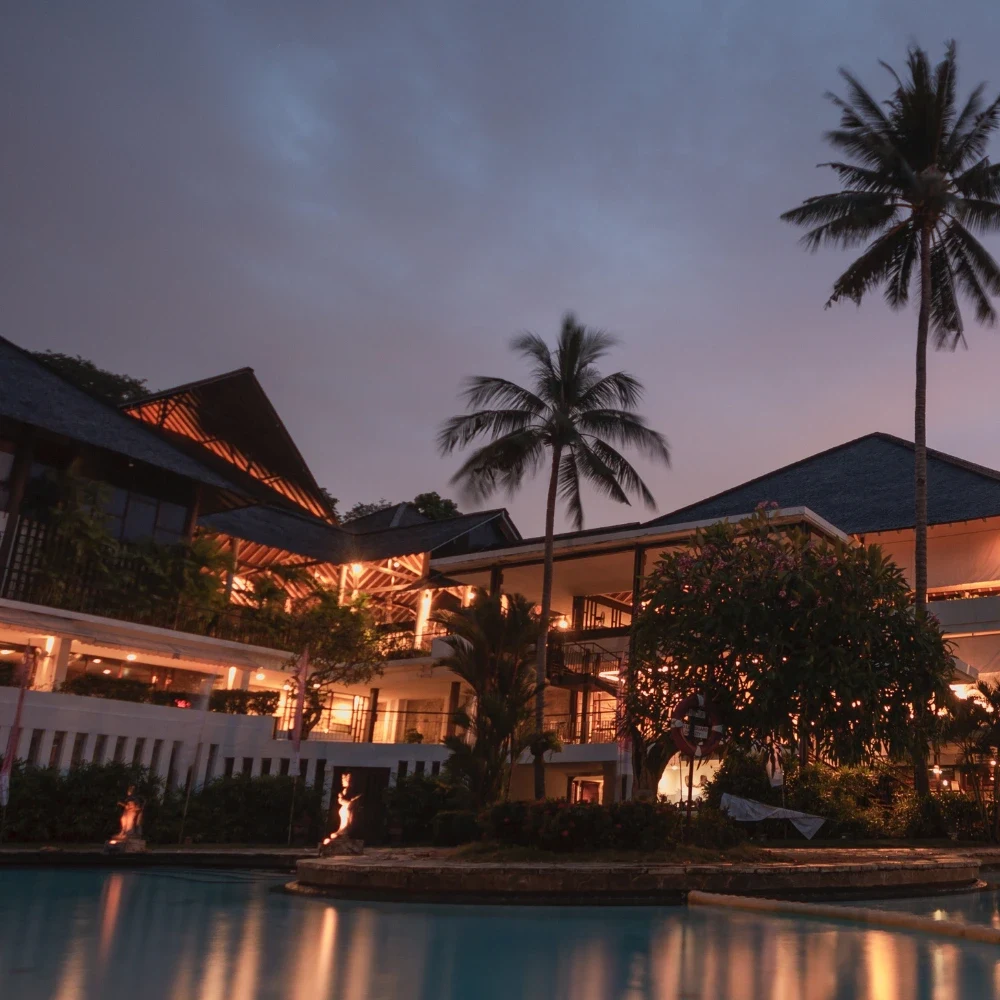 A lit resort building and palm trees reflected in a pool at dusk